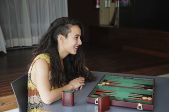 A woman with long dark hair smiles as she sits across a backgammon board in a cozy indoor setting