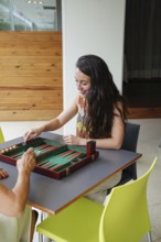 Mother and daughter are playing backgammon inside a cozy room The board is set on a table, and they
