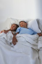 A young girl and boy, are captured sleeping peacefully side by side in a bright, cozy bed setting,