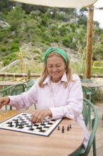 A senior woman plays chess outdoors on a sunny day, seated at a round table surrounded by greenery