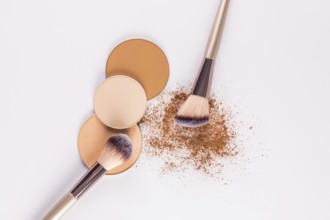 Top view of various shades of compact powders next to a makeup brush, with loose powder scattered