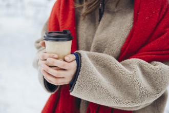 Cropped unrecognizable young woman in a red coat and cozy scarf holding a warm coffee cup outdoors
