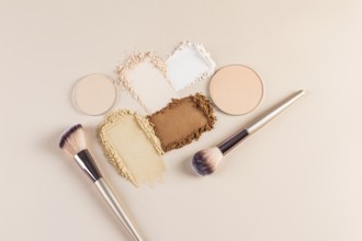 Top view of compact and loose face powders scattered artistically next to makeup brushes on a beige