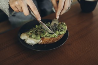 Close-up of hands using a fork and knife to slice avocado toast on a dark plate. The open-faced