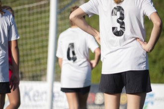 Female youth soccer players wearing white jerseys gather on the field near a goalpost. The sun