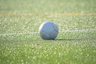 A soccer ball rests on a lush green soccer field, glistening under bright sunlight. The scene