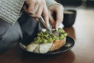 A close-up of hands slicing avocado toast topped with greens on a black plate, accompanied by a