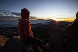 Back view of an unrecognizable man sitting on a mountain peak, watching a stunning sunrise over a