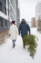 A couple walks hand in hand through a snow-covered urban area, carrying a freshly cut Christmas fir