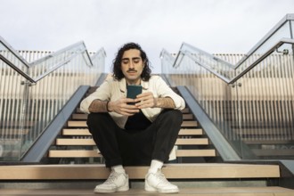 Young man sitting on outdoor stairs, casually browsing his smartphone The setting is urban and