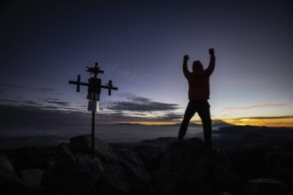 Unrecognizable man stands with arms raised near a silhouetted cross at sunrise, atop a rugged