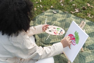 A black woman enjoys painting in a park, capturing creativity on canvas. Surrounded by a peaceful
