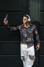 Stylish man wearing floral shirt and headphones takes a selfie. He wears sunglasses and ripped