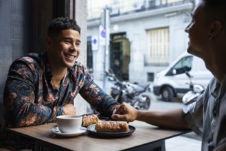 Two latin gay men hold hands, smiling and chatting over coffee and pastries in a warm, urban cafe