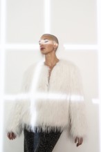 A middle-aged woman with a shaved head poses confidently, wearing a fashionable white faux fur coat