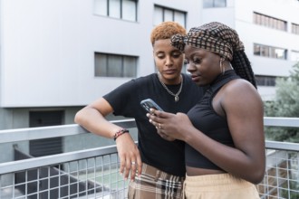 An LGBT multiethnic couple shares a tender moment outdoors, both focused on a smartphone, with one