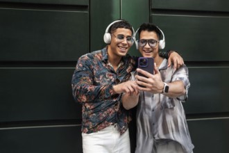 Two happy gay men wearing headphones, embracing and smiling while looking at a phone together.