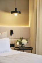 A cozy hotel room corner featuring a modern pendant lamp, cushioned headboard, and a side table