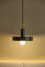 A modern hotel room showcases a sleek pendant light against a warm, neutral wall. The glowing bulb