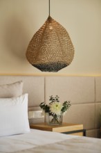 A cozy hotel room features a woven pendant lamp hanging above a wooden nightstand with a vase of