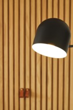 A modern hotel room featuring a sleek black lamp illuminating wooden panel walls. The warm lighting