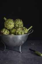 A collection of fresh artichokes sits in a rustic metal colander on a dark counter, accompanied by