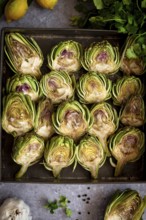 Top view of fresh artichoke halves on a baking tray, surrounded by lemons, herbs, and garlic,