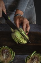 A person in a gray apron skillfully cuts an artichoke with a large kitchen knife on a dark wooden
