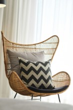 A stylish rattan chair adorned with cushy grey and black chevron pillows sits beside sheer