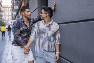 A smiling latin gay couple holds hands and enjoys a sunny day in the streets of Madrid, surrounded