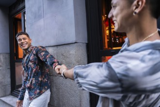 A smiling latin gay couple holds hands outside an urban cafe, radiating happiness and love. One