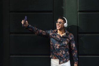 A young latin man in a floral shirt, wearing sunglasses and headphones, smiles while taking a