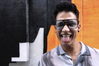 A young latin man wearing oversized sunglasses sticks out his tongue in a playful manner while