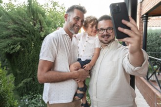 A joyful gay couple takes a smiling selfie with their son in a lush garden setting. The image