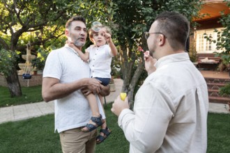 A gay couple enjoys a sunny afternoon in the garden with their young son, creating bubbles and