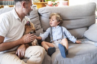 A joyful father and son duo share a warm moment with their small dog on a cozy sofa. The toddler,