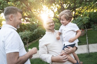 A joyful gay couple shares a tender moment with their son in a sunlit garden, surrounded by lush