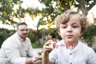 A young boy in a white shirt joyfully blows bubbles in a vibrant garden. His parent, watches in the