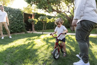 A joyful gay couple encourages their young son as he learns to ride a bicycle in a sunny garden.