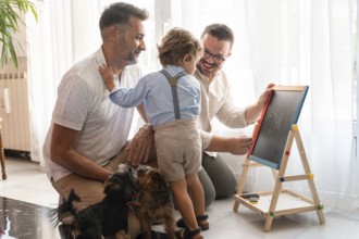 A joyful moment with a gay couple and their son drawing on a chalkboard, surrounded by playful dogs