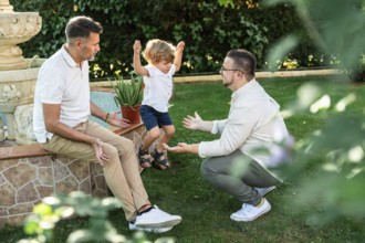 Joyful scene of a gay couple with their son in a lush garden. One father encourages their jumping
