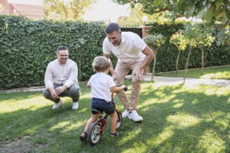 A joyful gay couple plays with their son, teaching him to ride a small bicycle in their garden. The