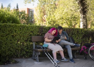 A couple studies together on a bench in a college garden The woman, with pink hair, uses a
