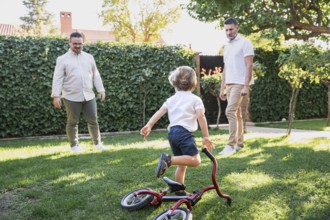 A loving gay couple shares a joyful moment with their young son playing with bicycle in the garden.