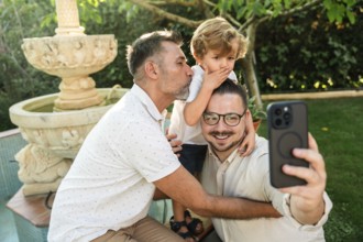 A joyful gay couple takes a selfie with their young son in a lush garden. One dad kisses the son