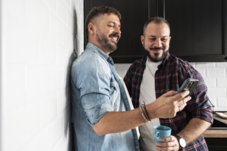 A gay couple enjoying a joyful moment in their kitchen while looking at a smartphone. One holds a