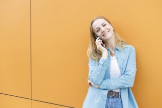 A cheerful woman stands against a bright orange wall in Warsaw, chatting on her phone She's dressed