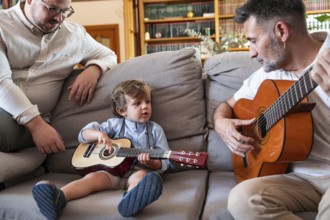 A loving gay couple sits on a sofa, teaching their young son to play the guitar. The warm,