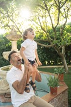 A father lifts his son as they gaze at something fascinating. Sunlight filters through the leaves