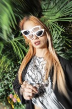 A fashionable woman with long hair wearing white cat-eye sunglasses and a stylish outfit stands